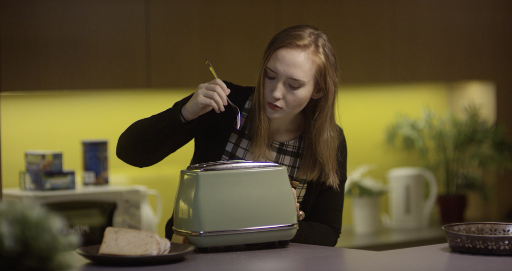 Woman with red hair in kitchen about to stick a fork into a green toaster. Still from video about quality Cybersecurity awareness training.
