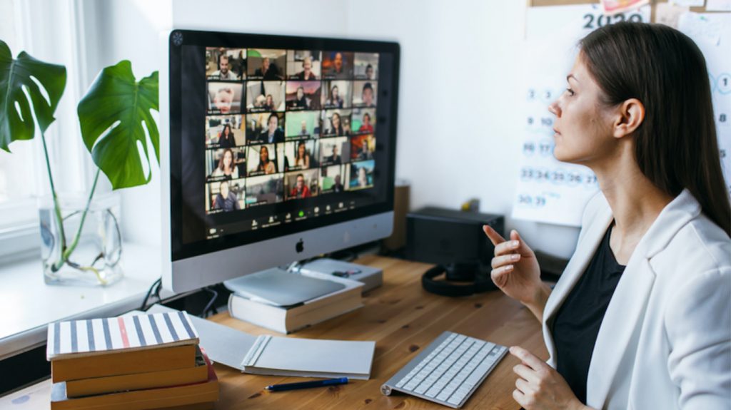 Woman working from home at an online meeting with multiple participants visible on computer screen. Indicating hybrid work and part of cybersecurity compliance training.